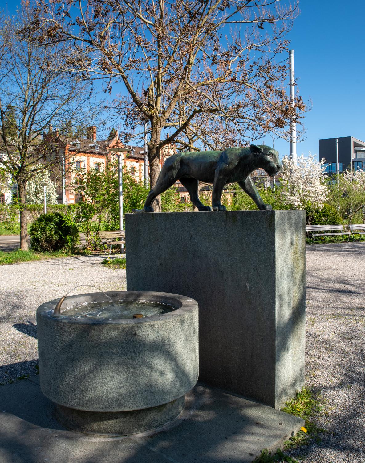 A round drinking fountain with, next to it, a bronze sculpture of a panther on top of a rectangular pedestal.