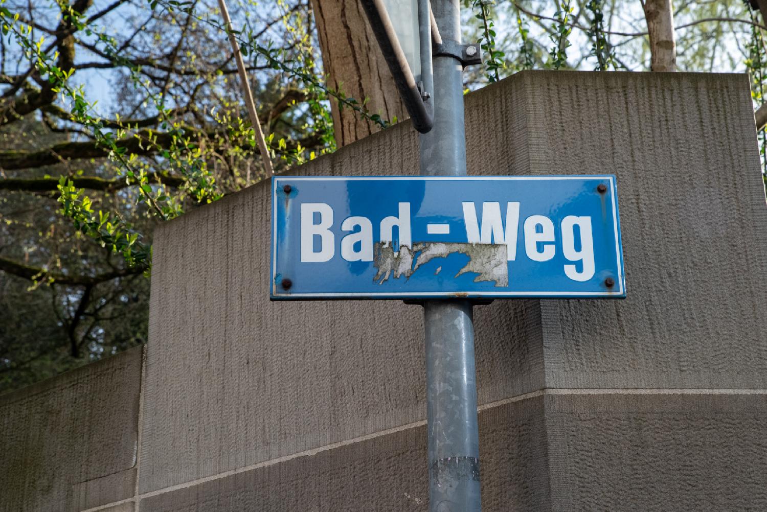 A blue "Bad-Weg" street sign on a metallic pole in front of a brown wall and some greenery. An old sticker has been badly removed from the sign.