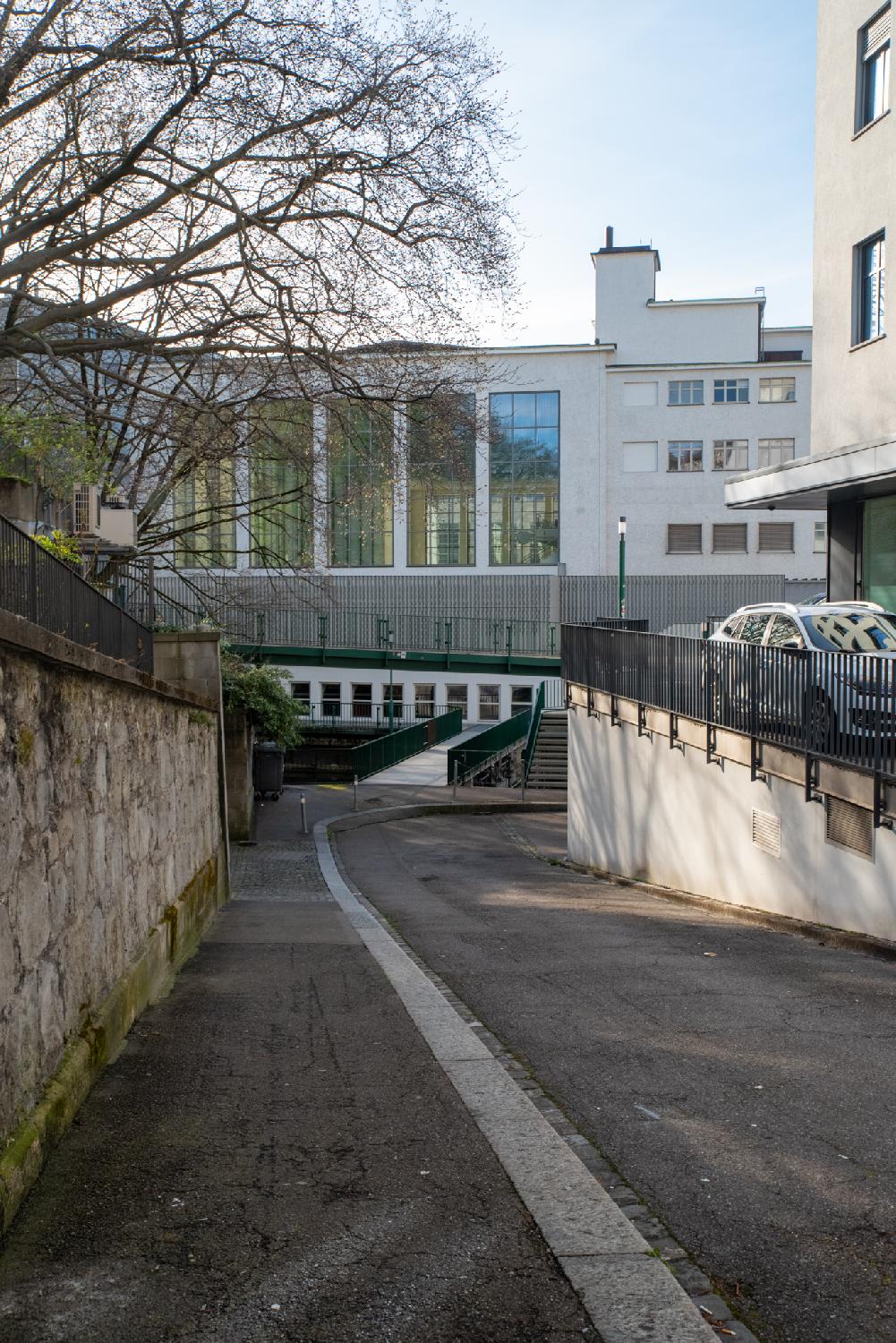 A narrow street going downhill, leading to a bridge and some stairs. In the background, a building with large windows reflects other buildings.