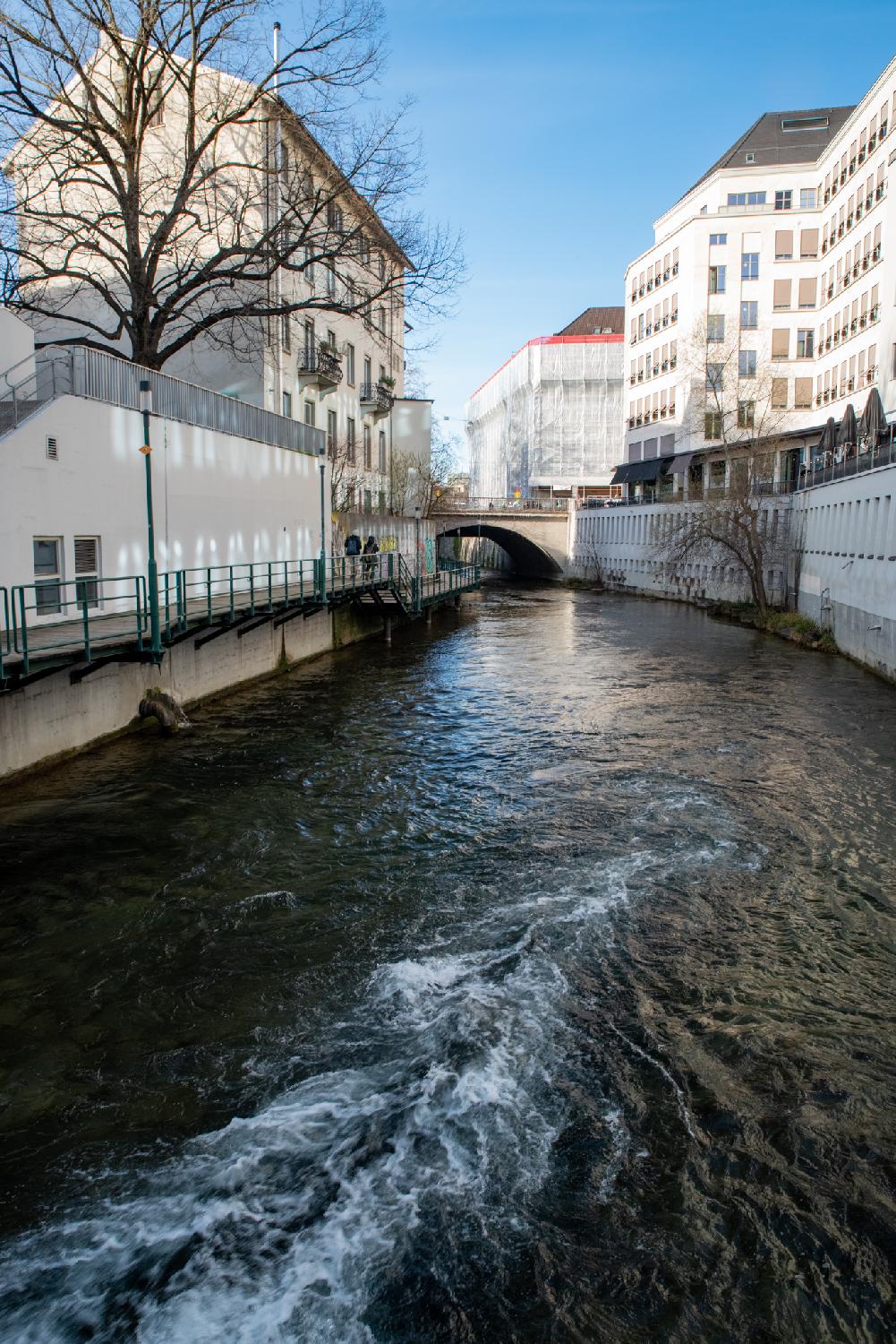 A dark canal between buildings, as seen from a bridge on top of it.