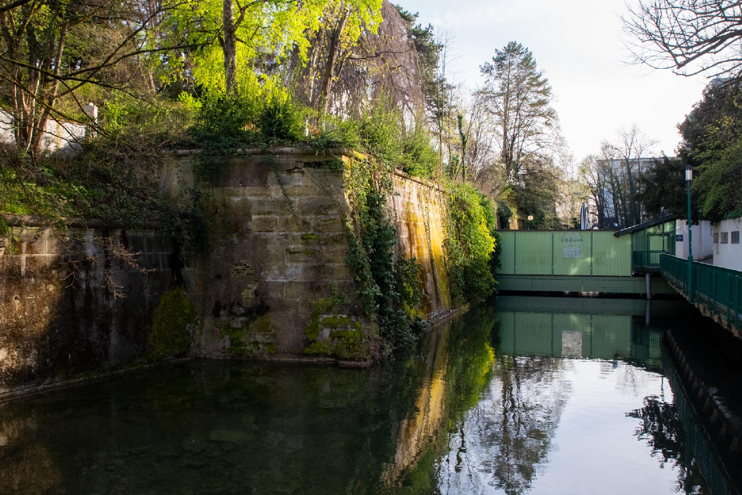 A canal between a stone wall covered with vegetation and trees and a green fence. The canal is blocked by a light green metallic wall with the logo for Rimini