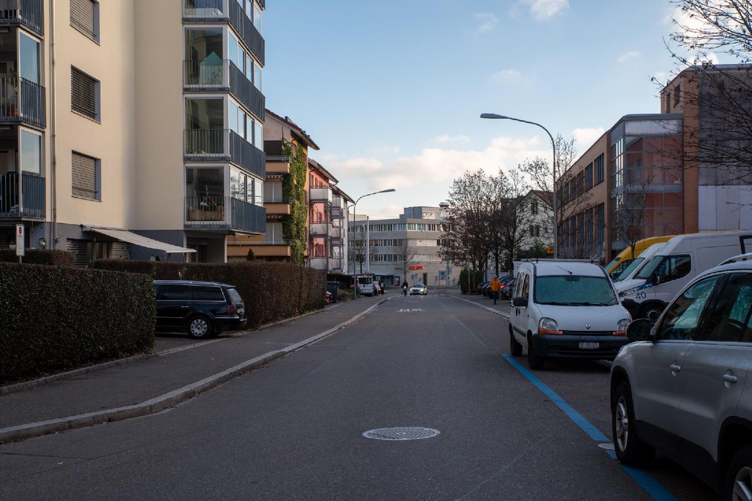 A street between 3-4-story residential buildings and a Migros supermarket in the background