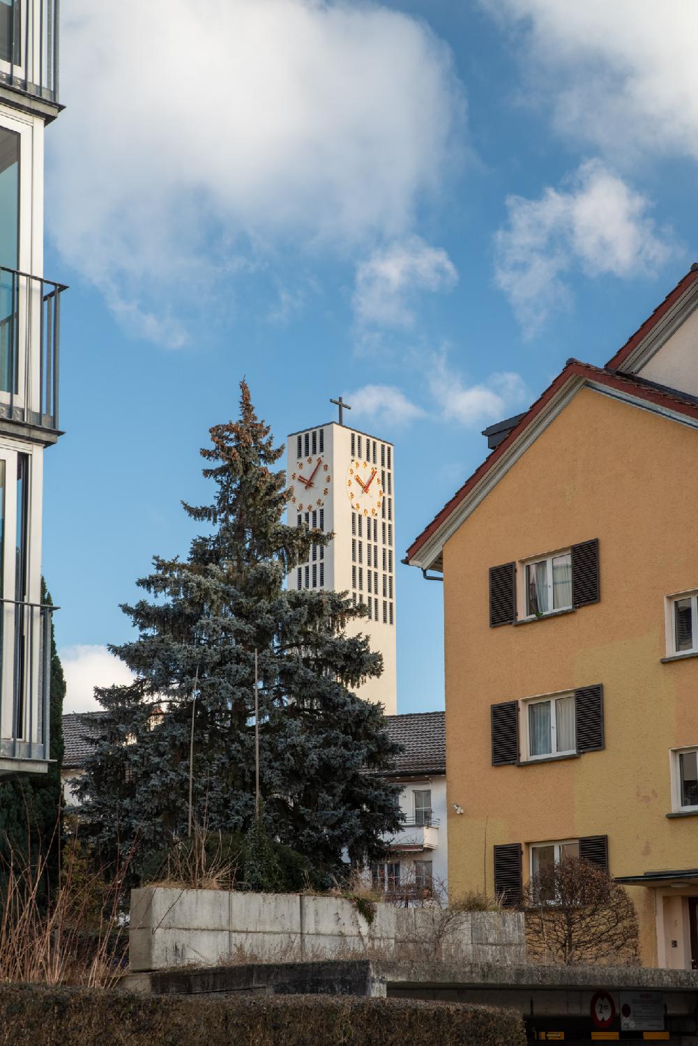 A white block church tower with a cross on top, behind a pine tree and between houses.