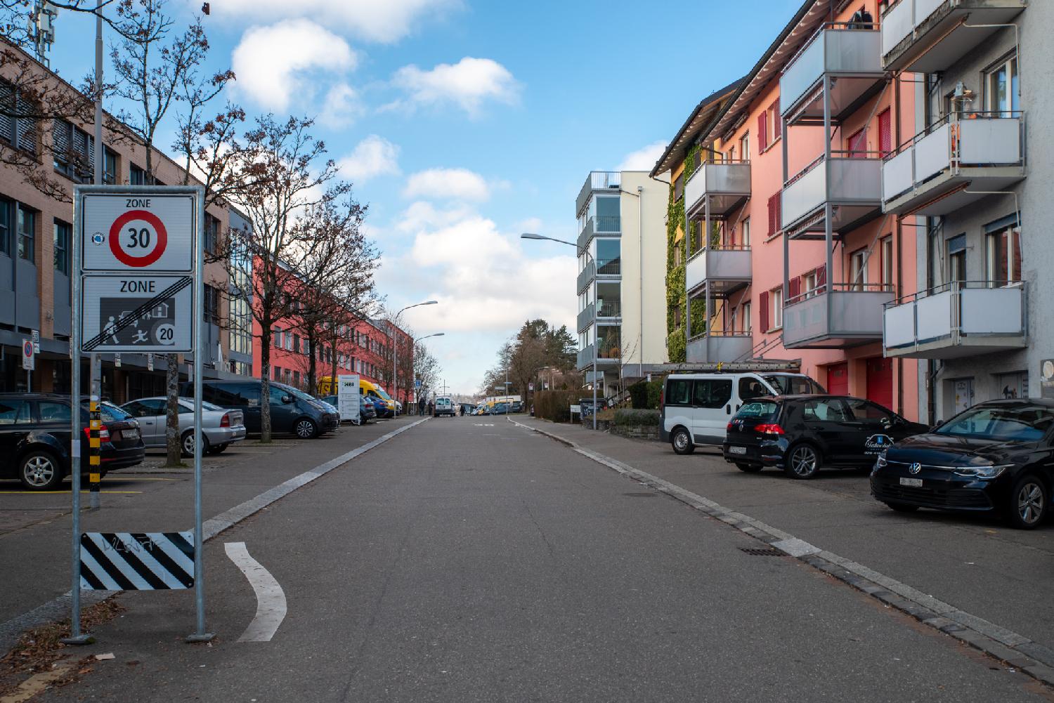 A residential street with a "Zone 30" sign in the foreground, 2-4-story buildings on each side and many cars parked on the sides.