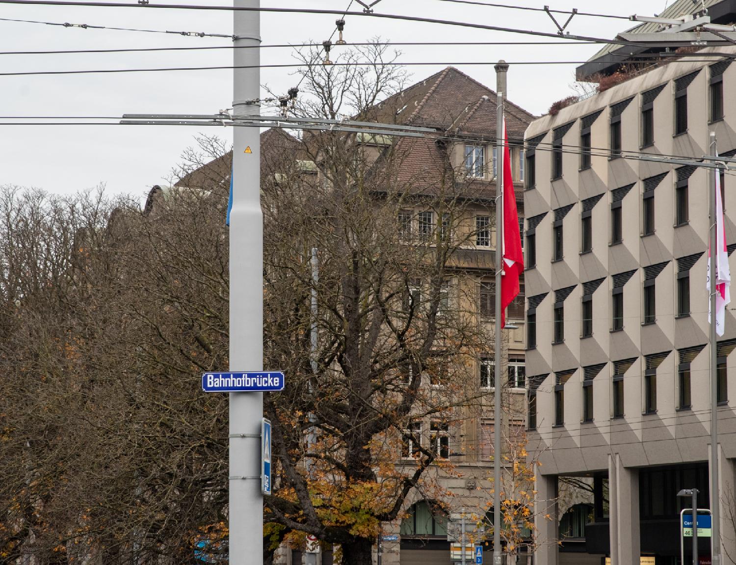 A blue "Bahnhofbrücke" street sign on a grey pole supporting tram cables. In the background: 5-story buildings, a tram stop, trees without leaves and a couple of red and white flags.