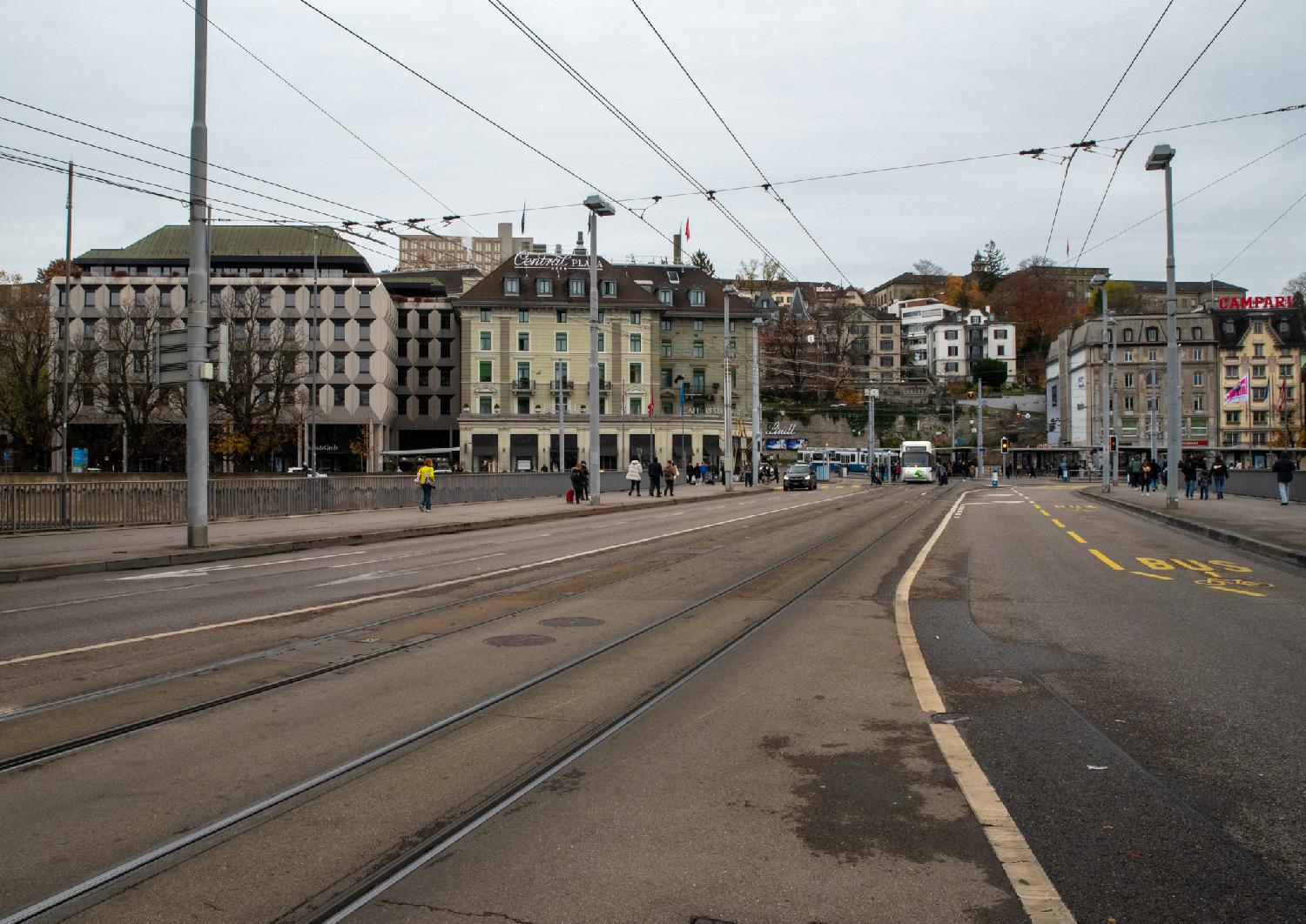 A wide bridge with tram rail tracks in the middle, two trams in the square in the middle, and 6-story city buildings in the background. The city in the background goes a bit uphill.
