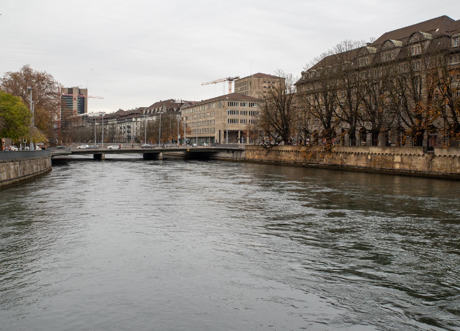 A river curving to the left, between stone walls, seen from a bridge. The right bank of the river is filled with 5-6-story buildings and leafless trees. Another bridge with cars is visible not very far from the photographer's point of view.