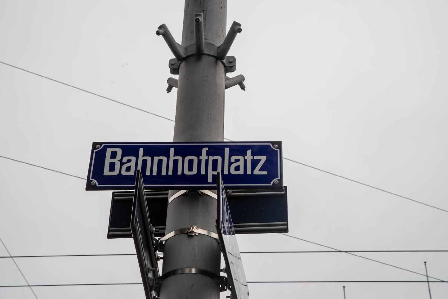 A grey pole with blue street plates on it; the one visible shows Bahnhofplatz. There are tram electrity cables in the background.