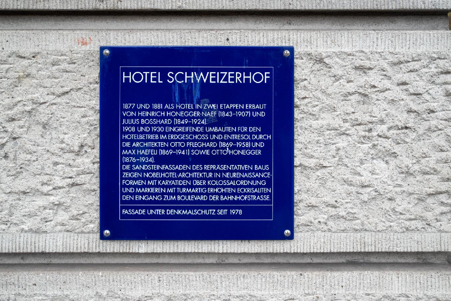 A blue plate with a text in German reading "Hotel Schweizerhof. 1877 und 1881 als Hotel in zwei Etappen erbaut von Heinrich Honegger-Naef (1843-1907) und Julius Bosshard (1849-1924). 1908 und 1930 eingreifende Umbauten für den Hotelbetrieb im Erdgeschoss und Entresol durch die Architekten Otto Pfleghard (1869-1958) und Max Haefeli (1869-1941) sowie Otto Honegger (1876-1934). Die Sandsteinfassaden des repräsentativen Baus zeigen Nobelhotel-Architektur in Neurenaissanceformen mit Karyatiden über Kolossarordnung und markieren mit tormartig eröhten Eckrisaliten den Eingang zum Boulevard der Bahnhofstrasse.Fassade under Denkmalschutz seit 1978"