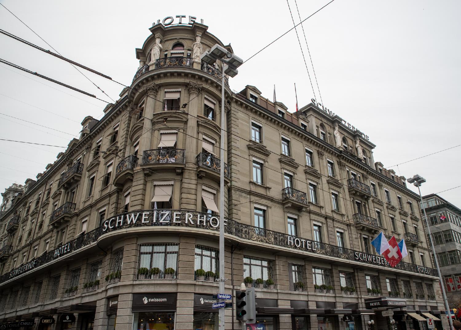 A large sandstone corner building, with balconies on the second floor lettered Hotel Schweizerhof and, in the ground floor, a hoter and multiple shops. The corner is round and has columns and statues.