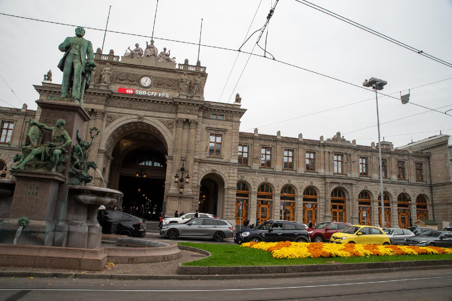 Façade of the Hauptbahnhof in Zürich, a large portal with the SBB logo on top, and a row of arched windows next to it. In front of the building, there's a fountain statue of a man, and a parterre of yellow and orange flowers. A row of cars is between the building and the foreground elements.
