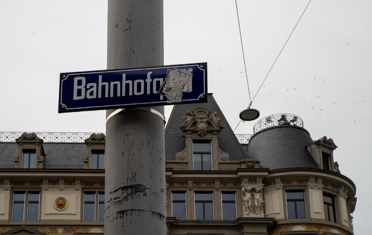 A metallic pole with a blue sign for Bahnhofquai. An old sticker hides the "quai" part of the name. In the background, buildings with dormer windows.