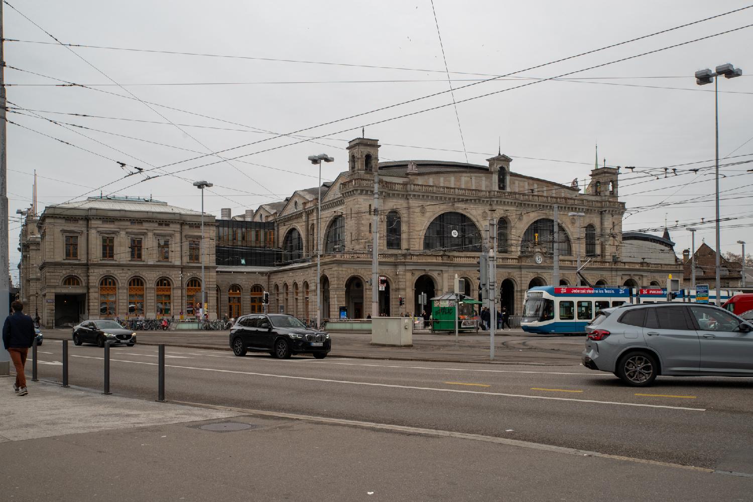 A large train statino building with a visible aisle. There are arched windows and doors, and small towers in the corners. The street in front of it has a tram and a few cars on the road.