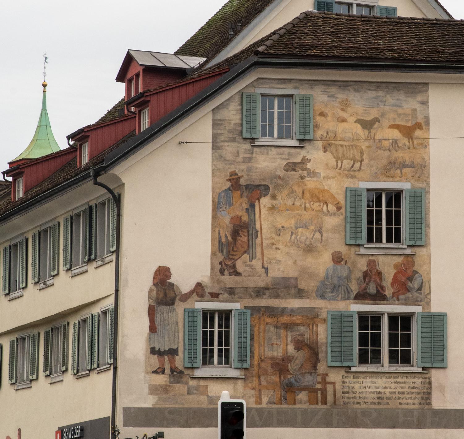 A mural painted on a wall. The wall also has four windows with blue blinds. The painting represents people, including someone using a loom, and sheep.