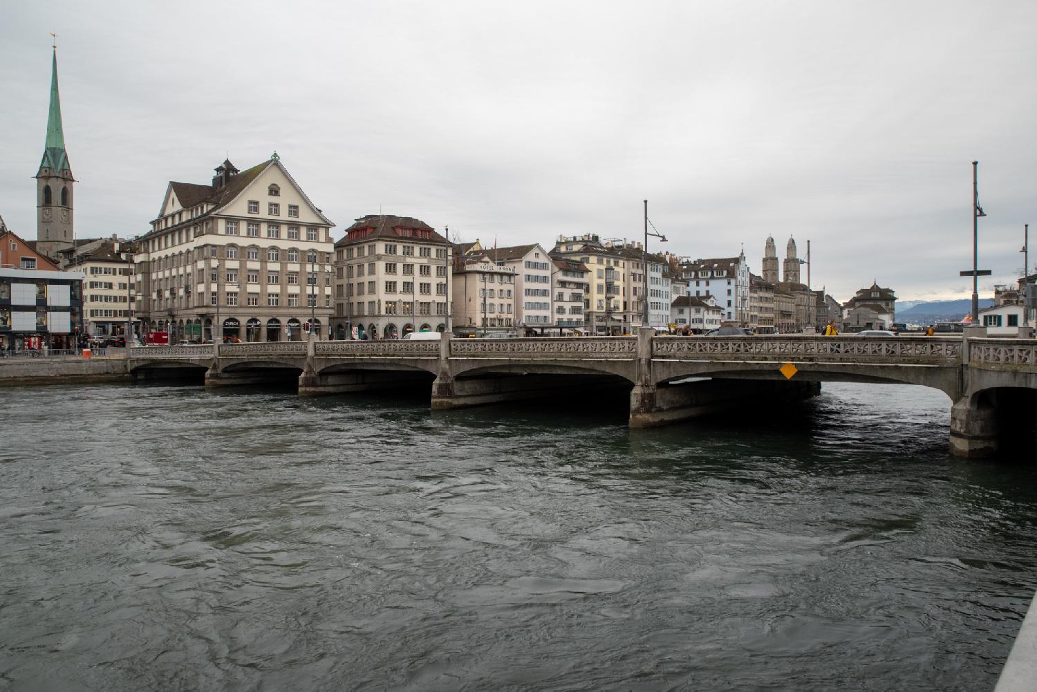 A river crossed by a stone bridge, with two churches and multiple 5-6 story buildings visibles on the other side of the river