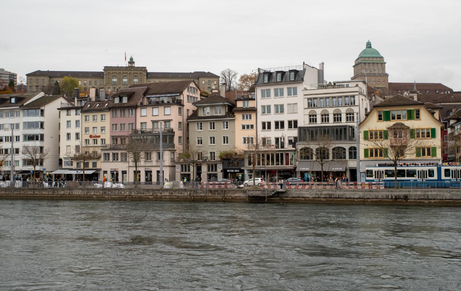 Multiple 5-6 story buildings, seen from across a river. Two large buildings are also visible on a higher ground above the foreground buildings.