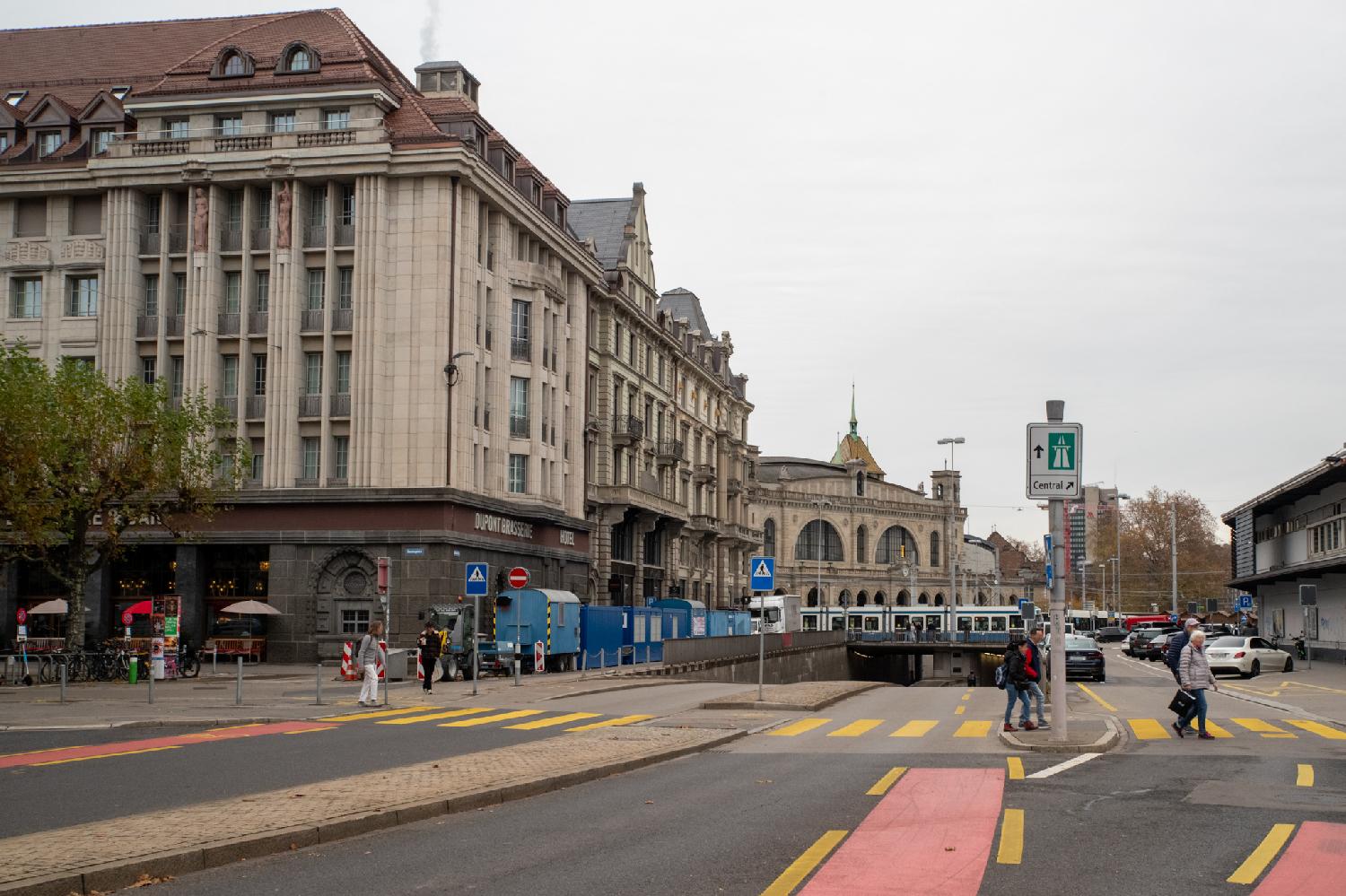 A street that splits between an underground tunnel and a surface area, with pedestrian zebras; in the background, the building of the Hauptbahnhof; on the left side of the street, imposing 6-story buildings.