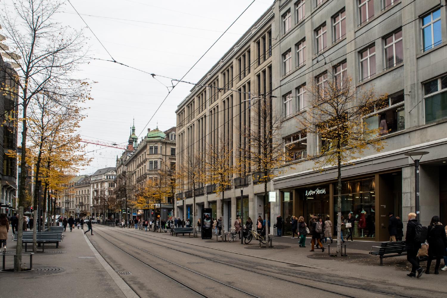 A wide street with tramway tracks, wide sidewalks, and 5-6-story buildings on each sides, with shops at the ground floor