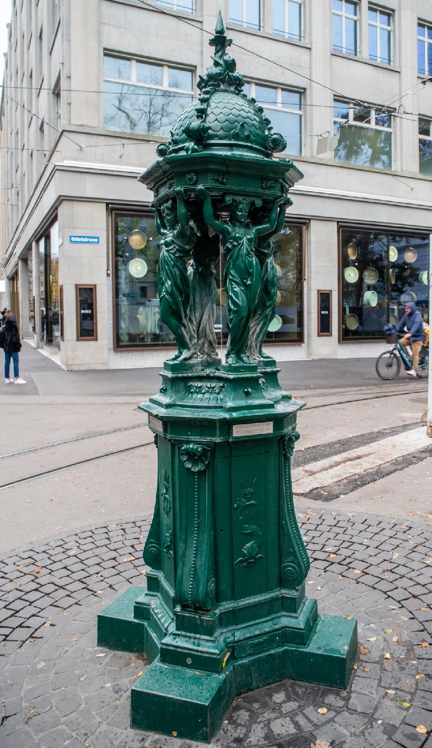 A green cast iron fountain with a high base and water flowing vertically between four caryatids holding a domed roof