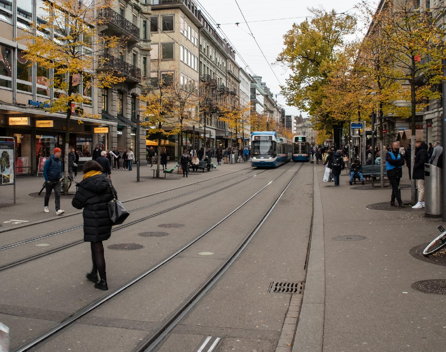 A street with two train tracks and a tram on each one, and wide sidewalks with people standing and sitting, between 5-story buildings