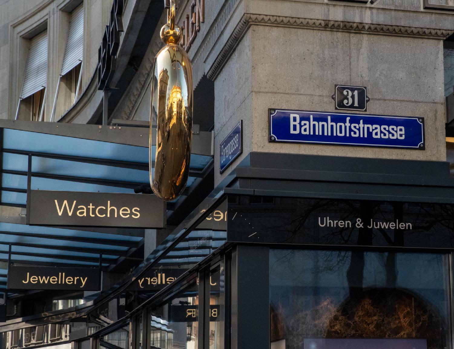 A blue Bahnhofstrasse sign on a wall, on top of a jewlery shop that has a golden gigantic watch as a sign which reflects the building behind it