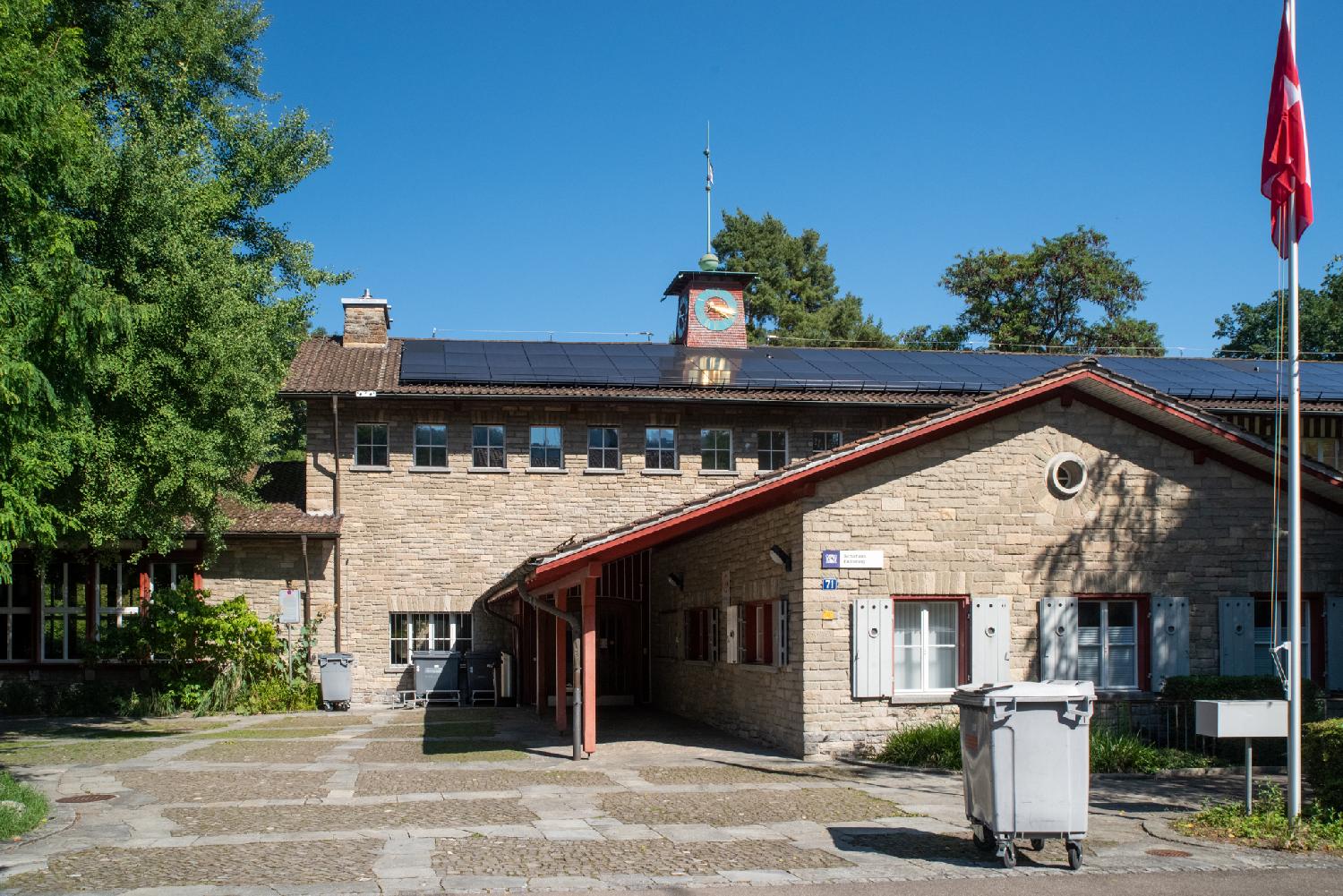 Some connected building forming a larger structure. The largest building is long building with a row of windows on top, solar panels on the roof, and a small clock tower on the roof. There's a mast with a Swiss flag and a large trash container in the foreground.