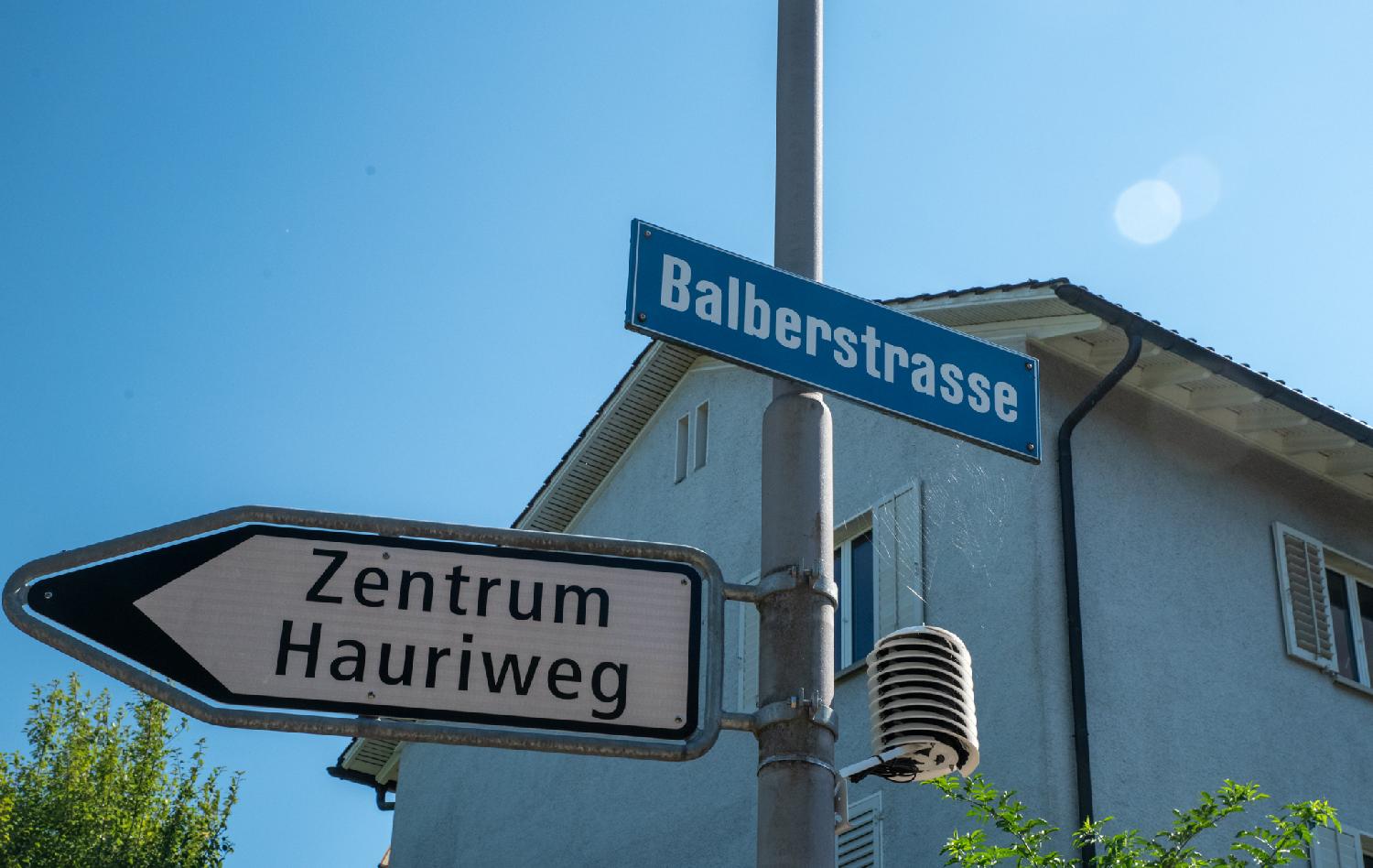 A metallic pole with an arrow pointing left to Zentrum Hauriweg, and a blue Balberstrasse street sign. There's also some kind of small spirally measurement device, and a spider made a web between that and the street sign. There's a residential building in the background.