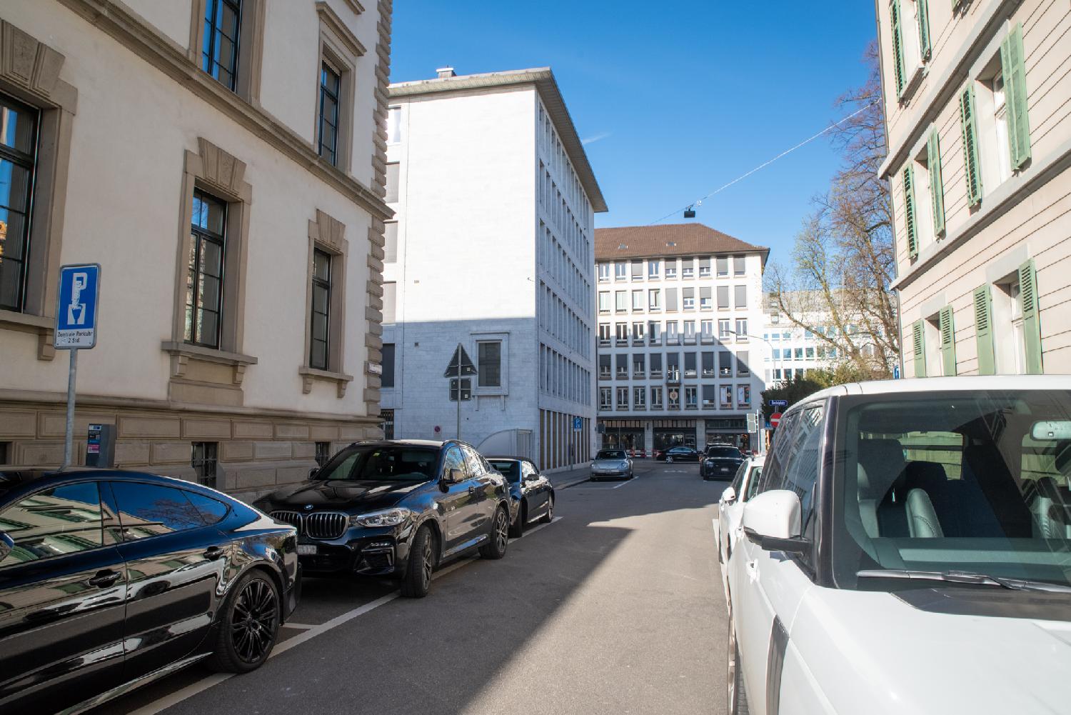 A narrow, one-way street with large cars parked on both sides, between 6-story buildings.