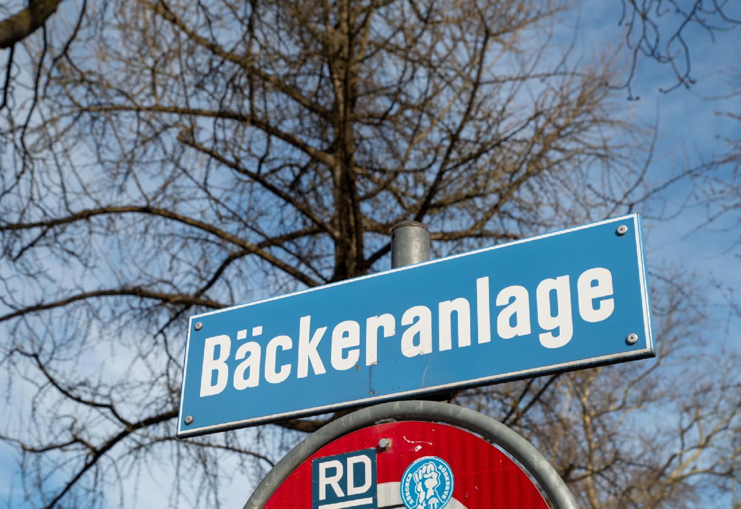 A blue Bäckeranlage street sign, in front of a blurred tree and a blue sky