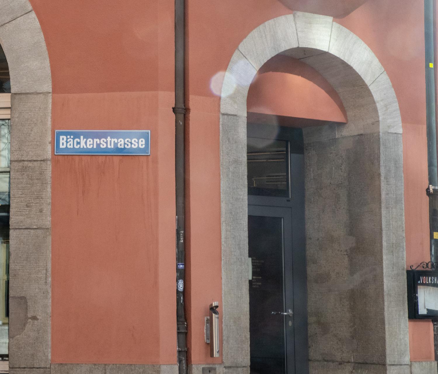 A blue Bäckerstasse sign on a brick-colored painted wall next to an arched door