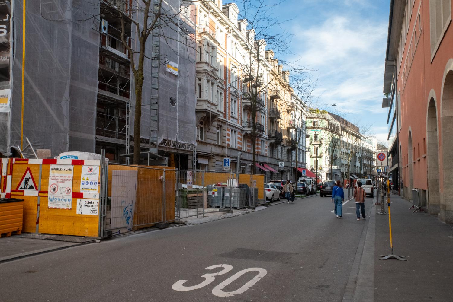 A street with a 30 painted on the ground between rows of 6-stories residential buildings. In the foreground, the building is hidden behind scaffolding.