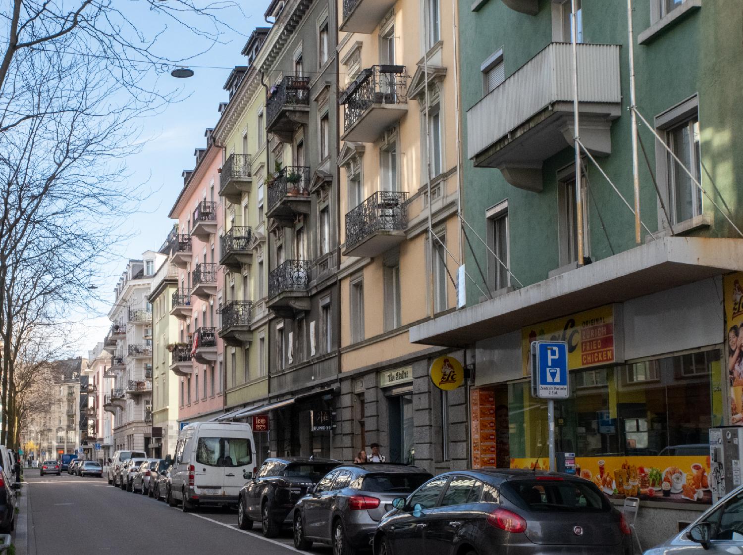 A street with 6-stories pastel-colored façades on the right and a row of cars on each side