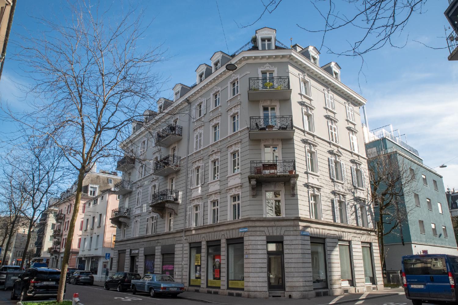 A grey 6-storey residential building on the corner of a street with dormer windows and metallic balconies