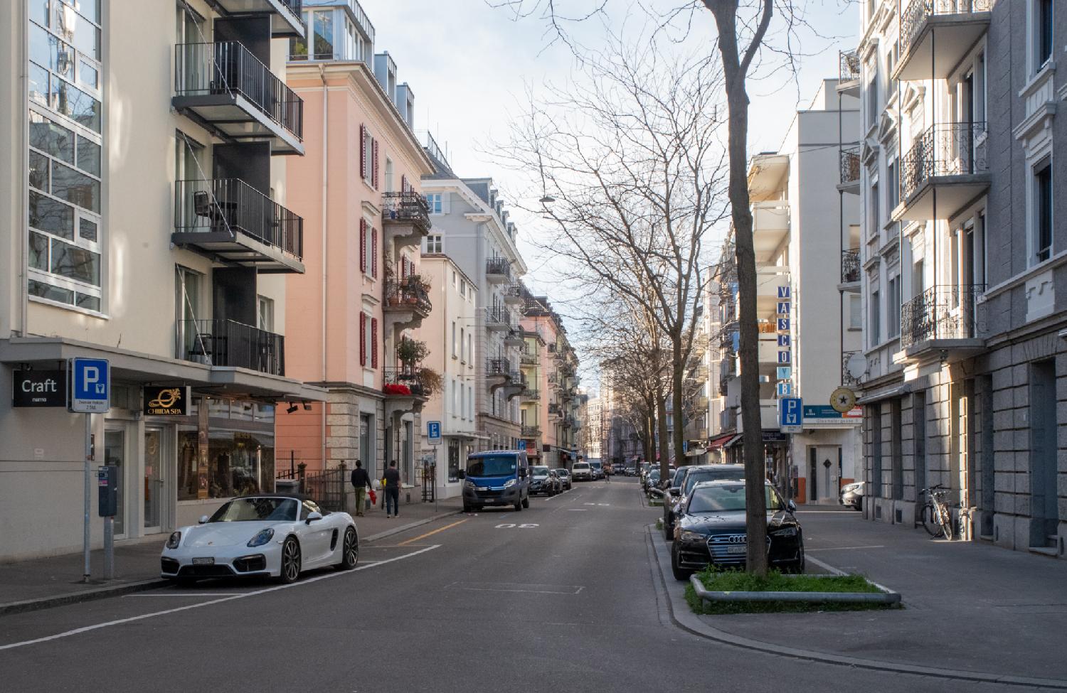 A street between 5-6-stories residential buildings, a row of trees on the right and cars parked on each side